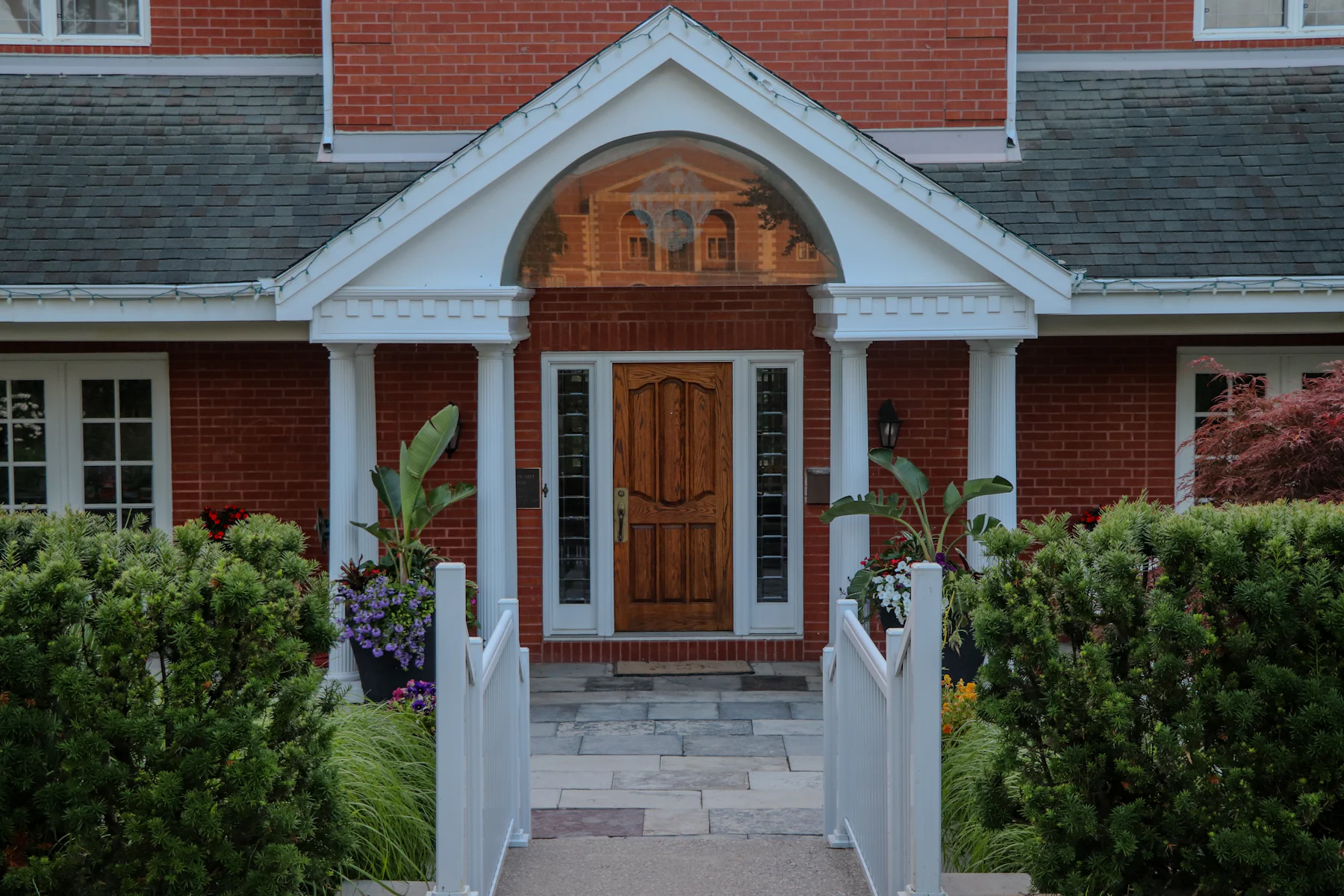The front entrance of a red brick house: a white arched portico over a wooden door, with steps and clipped shrubs on either side.