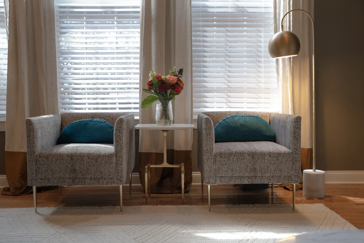 Two grey armchairs with teal cushions either side of a small table with flowers, in front of two blinded windows and a brass floor lamp.