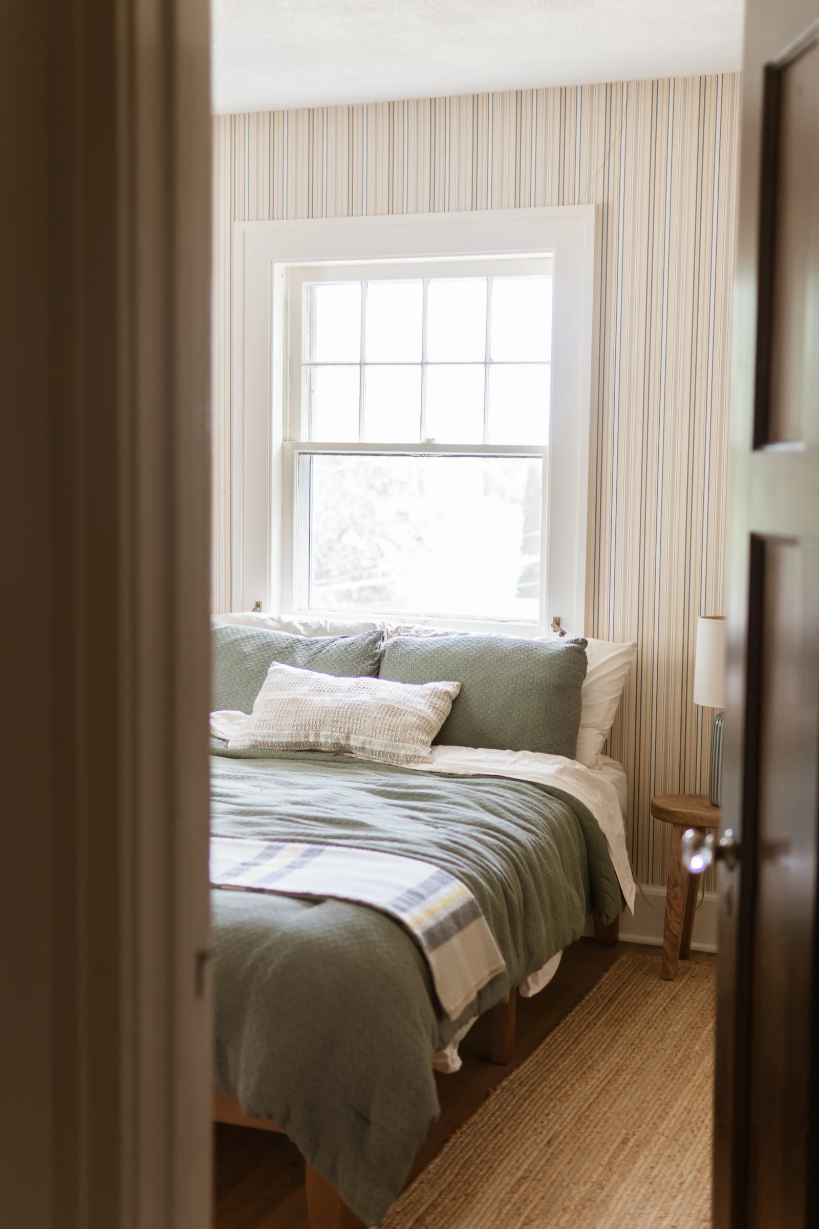 Seen through an open doorway, a bed with a soft green cover and a striped blanket sits under a bright sash window, against striped wallpaper, with a wooden stool and a lamp beside it.