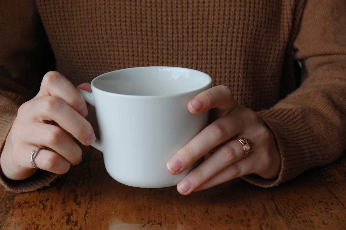Two hands in a brown sweater hold a white mug on a worn wooden table.