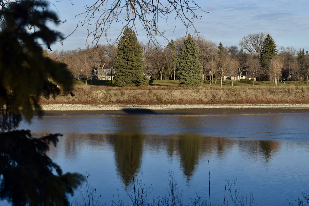 A wide, calm river in late autumn reflecting a blue sky, with clay banks, spruce, bare elms, and houses along the far shore.
