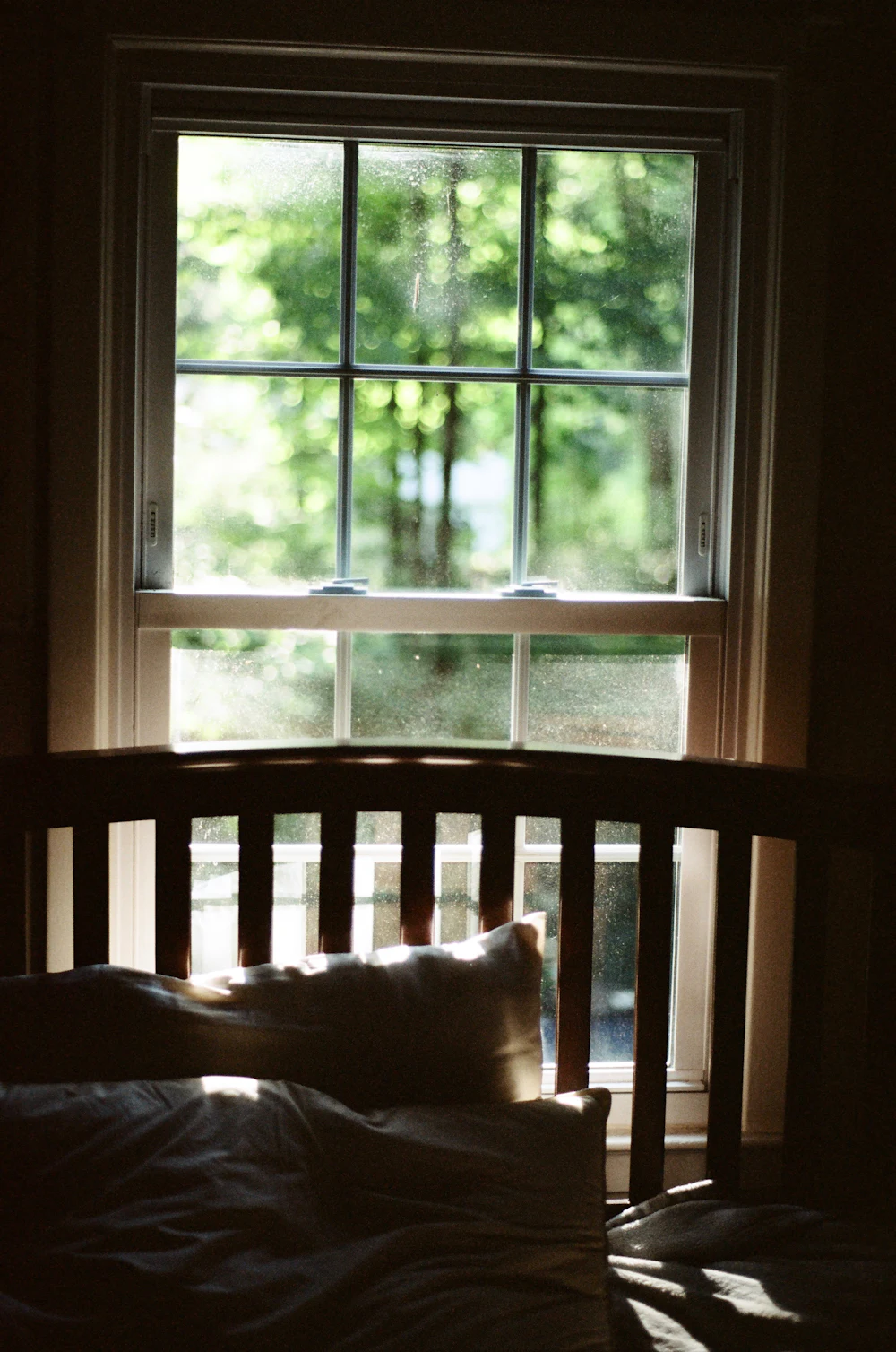 A sash window looking onto green trees, with the slatted wooden headboard of a bed and its pillows in the dark foreground.