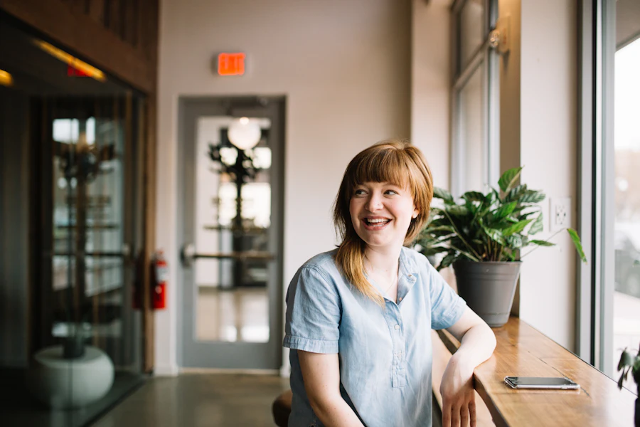 A woman with red hair in a pale blue shirt leans on a counter by a tall window, smiling at something off to one side.
