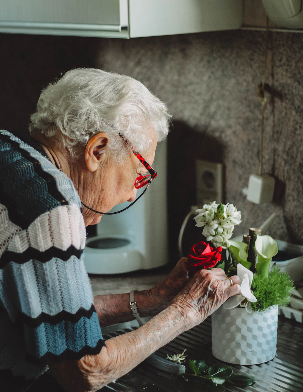An older woman in a striped cardigan bends over a small pot of flowers at a kitchen counter.