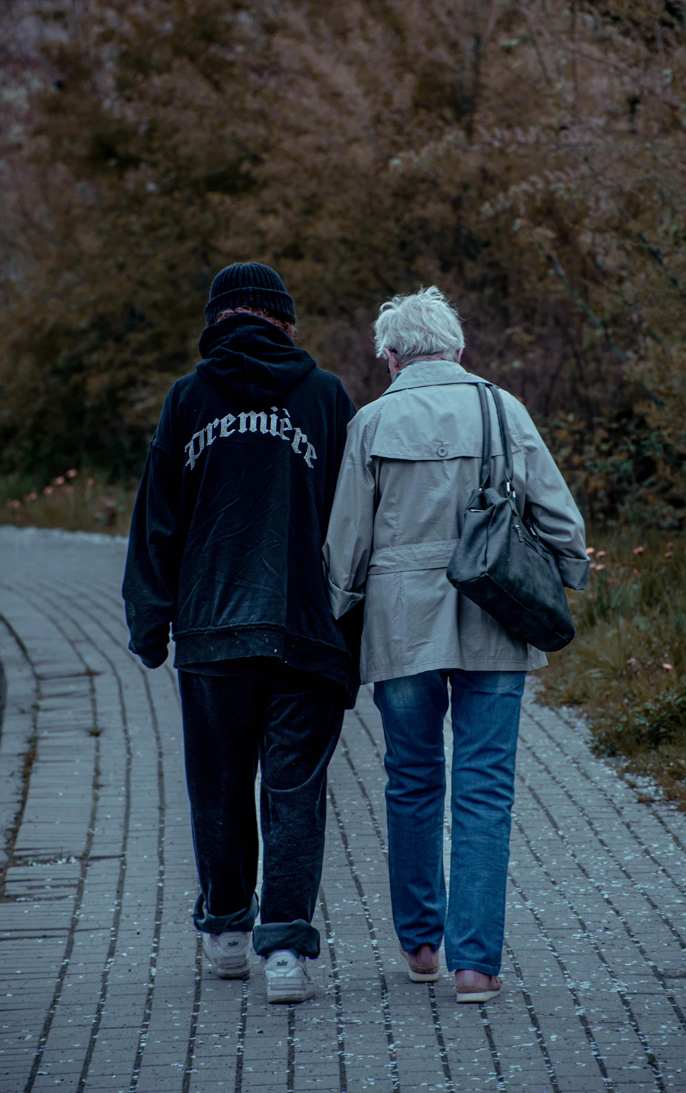 Two people walk away along a paved path, one in a dark hoodie and one in a grey jacket.