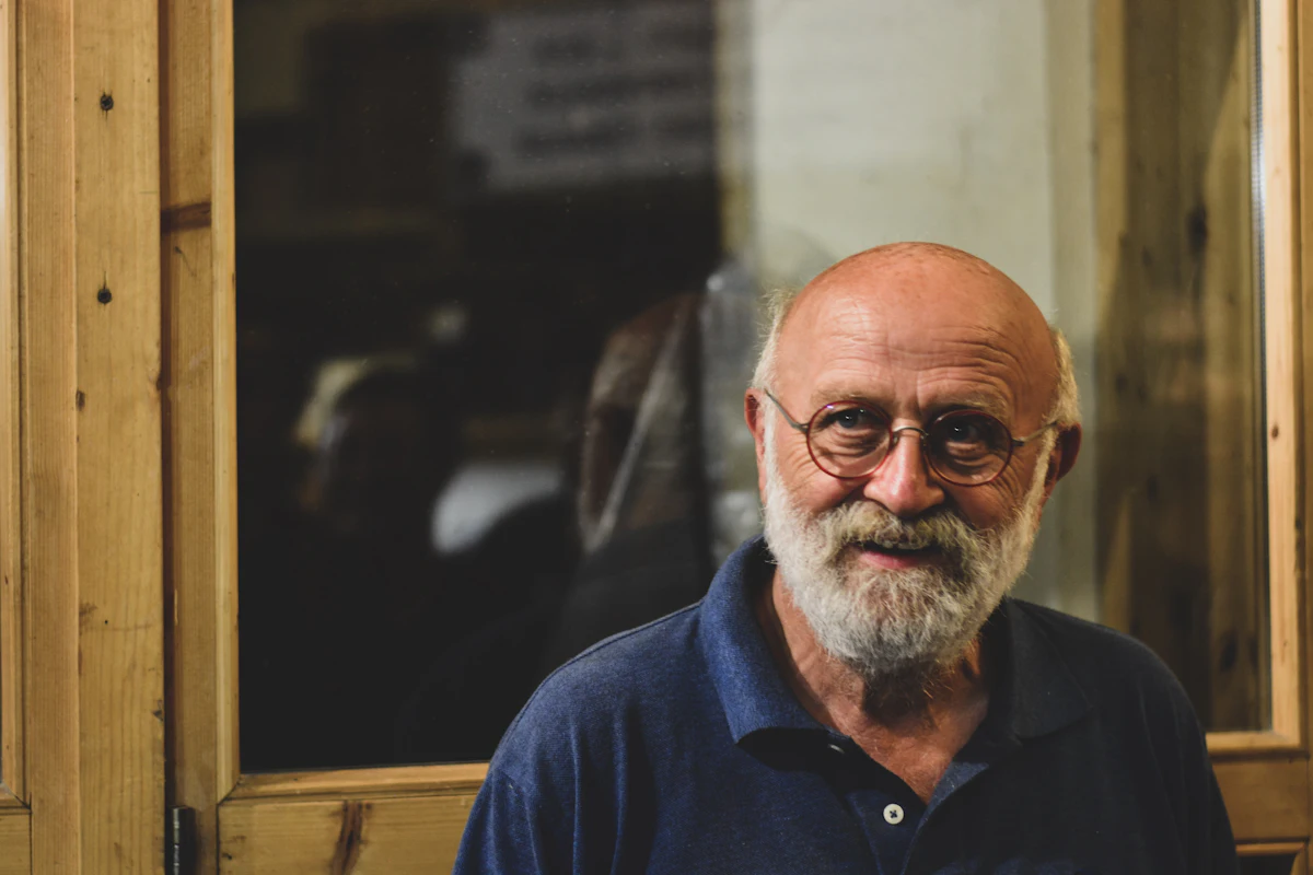 An older man with a white beard and glasses, in a navy shirt, smiles slightly as he stands in front of a wood-framed window.